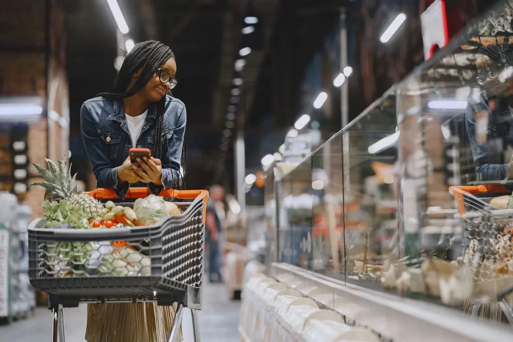 A shopper with a trolley checking an order on her phone in a supermarket aisle